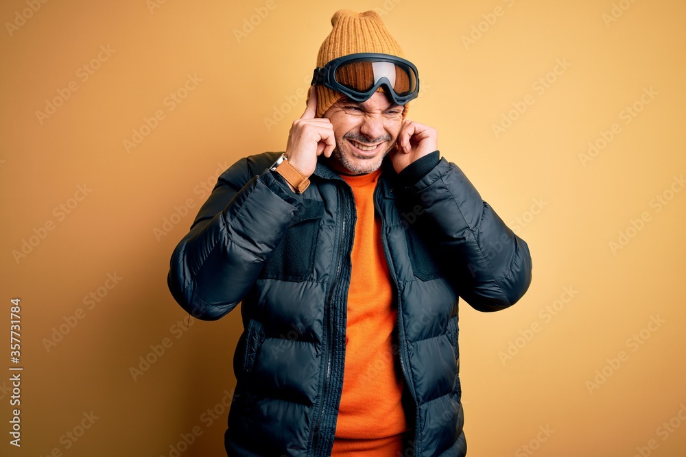Young handsome skier man skiing wearing snow sportswear using ski goggles covering ears with fingers with annoyed expression for the noise of loud music. Deaf concept. Young handsome skier man skiing wearing snow sportswear using ski goggles covering ears with fingers with annoyed expression for the noise of loud music. Deaf concept.