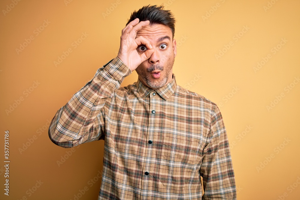 Young handsome man wearing casual shirt standing over isolated yellow background doing ok gesture shocked with surprised face, eye looking through fingers. Unbelieving expression. Young handsome man wearing casual shirt standing over isolated yellow background doing ok gesture shocked with surprised face, eye looking through fingers. Unbelieving expression.