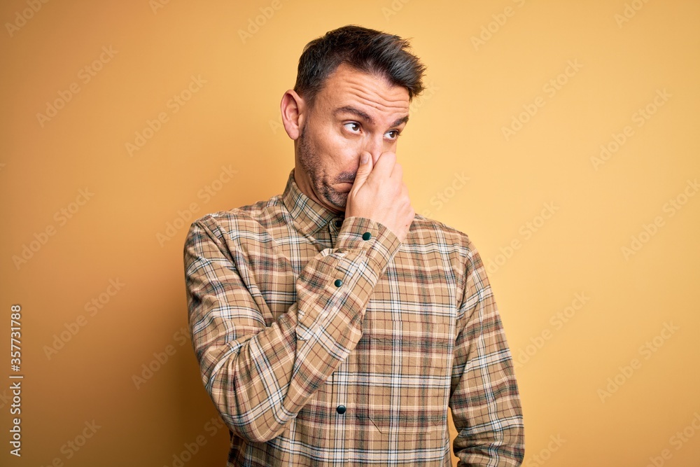 Young handsome man wearing casual shirt standing over isolated yellow background smelling something stinky and disgusting, intolerable smell, holding breath with fingers on nose. Bad smell Young handsome man wearing casual shirt standing over isolated yellow background smelling something stinky and disgusting, intolerable smell, holding breath with fingers on nose. Bad smell