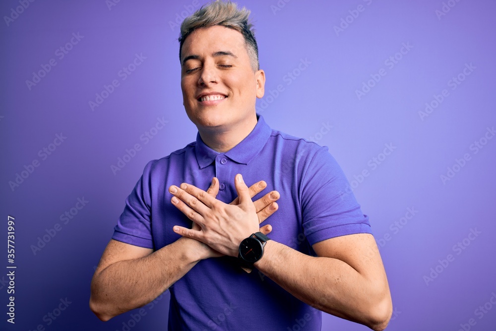 Young handsome modern man wearing casual purple t-shirt over isolated background smiling with hands on chest with closed eyes and grateful gesture on face. Health concept. Young handsome modern man wearing casual purple t-shirt over isolated background smiling with hands on chest with closed eyes and grateful gesture on face. Health concept.