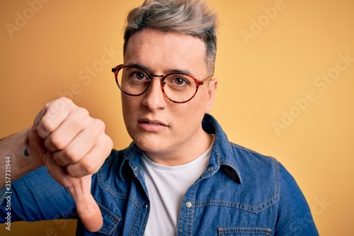 Close up of young handsome modern man wearing glasses and denim jacket over yellow background with angry face, negative sign showing dislike with thumbs down, rejection concept