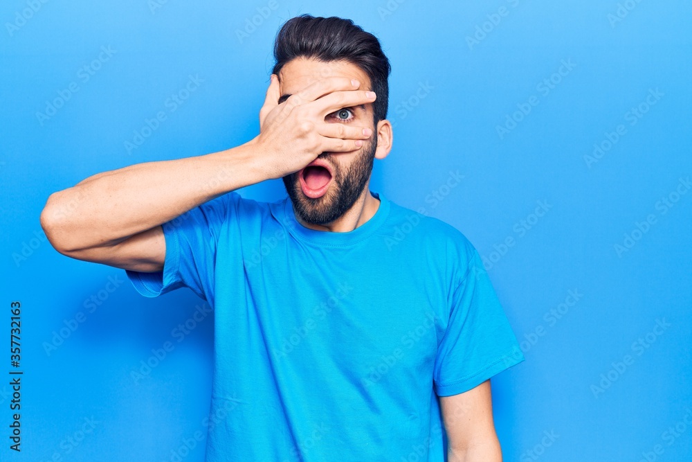 Young handsome man with beard wearing casual t-shirt peeking in shock covering face and eyes with hand, looking through fingers afraid Young handsome man with beard wearing casual t-shirt peeking in shock covering face and eyes with hand, looking through fingers afraid