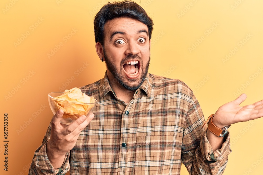 Young hispanic man holding potato chip celebrating achievement with happy smile and winner expression with raised hand Young hispanic man holding potato chip celebrating achievement with happy smile and winner expression with raised hand