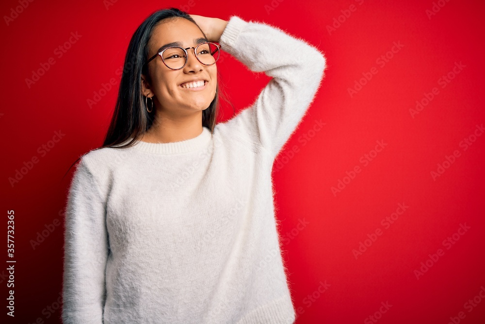 Young beautiful asian woman wearing casual sweater and glasses over red background smiling confident touching hair with hand up gesture, posing attractive and fashionable