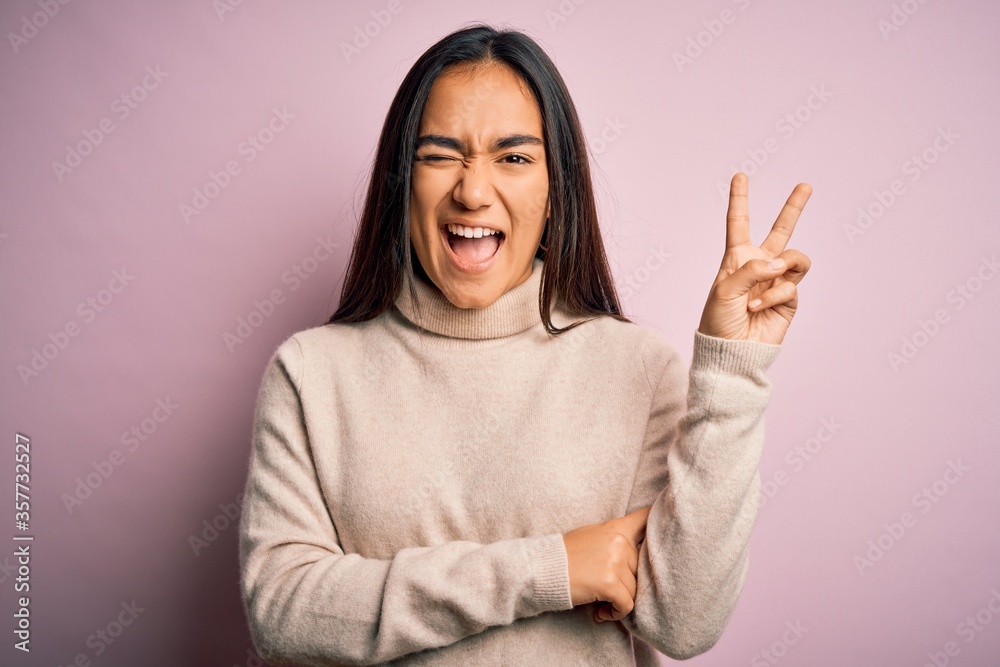Young beautiful asian woman wearing casual turtleneck sweater over pink background smiling with happy face winking at the camera doing victory sign. Number two. Young beautiful asian woman wearing casual turtleneck sweater over pink background smiling with happy face winking at the camera doing victory sign. Number two.