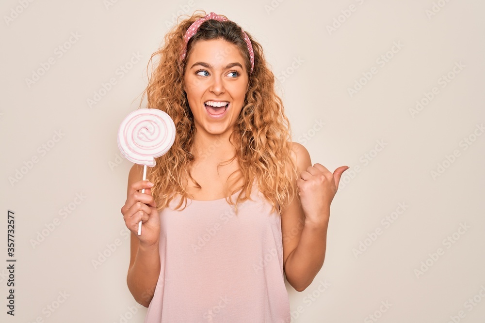 Young beautiful blonde woman with blue eyes eating sweet candy lollipop over pink background pointing and showing with thumb up to the side with happy face smiling Young beautiful blonde woman with blue eyes eating sweet candy lollipop over pink background pointing and showing with thumb up to the side with happy face smiling