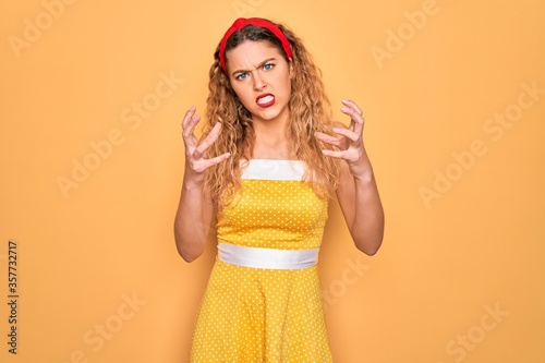 Beautiful blonde pin-up woman with blue eyes wearing diadem standing over yellow background Shouting frustrated with rage, hands trying to strangle, yelling mad