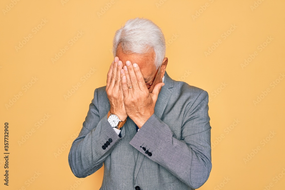 Middle age senior grey-haired handsome business man wearing glasses over yellow background with sad expression covering face with hands while crying. Depression concept.