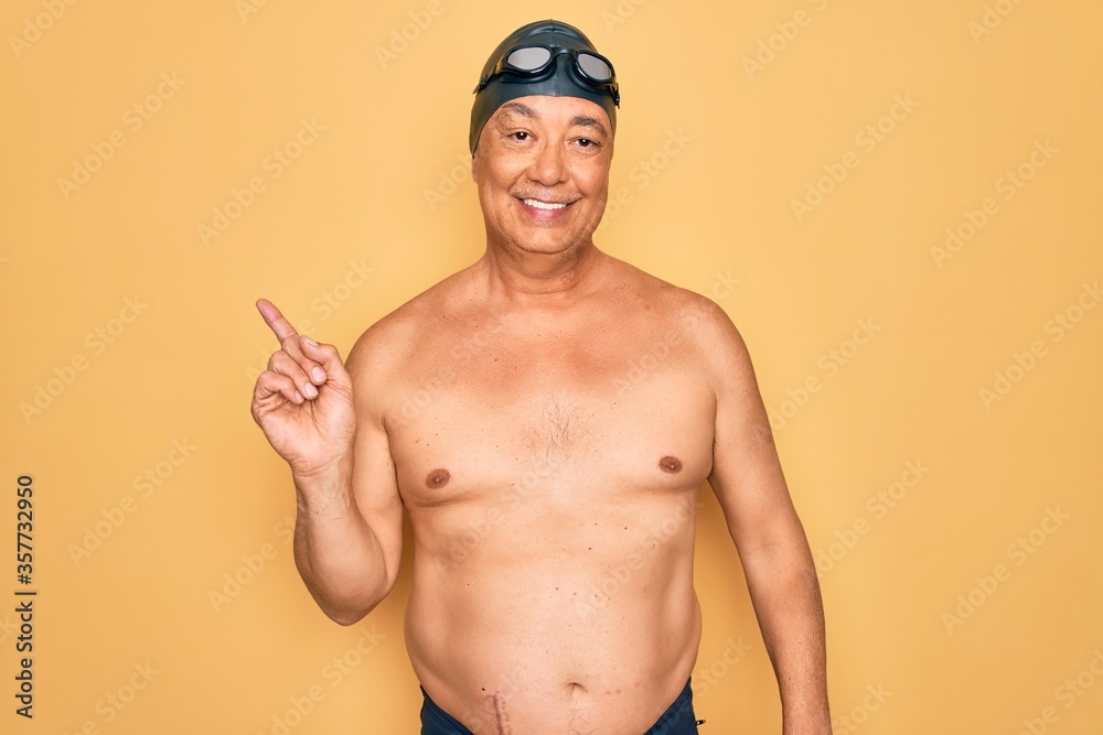 Middle age senior grey-haired swimmer man wearing swimsuit, cap and goggles with a big smile on face, pointing with hand finger to the side looking at the camera.