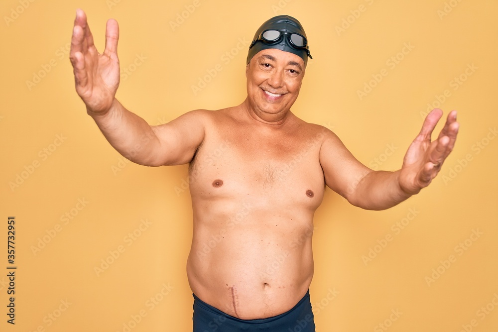 Middle age senior grey-haired swimmer man wearing swimsuit, cap and goggles looking at the camera smiling with open arms for hug. Cheerful expression embracing happiness.