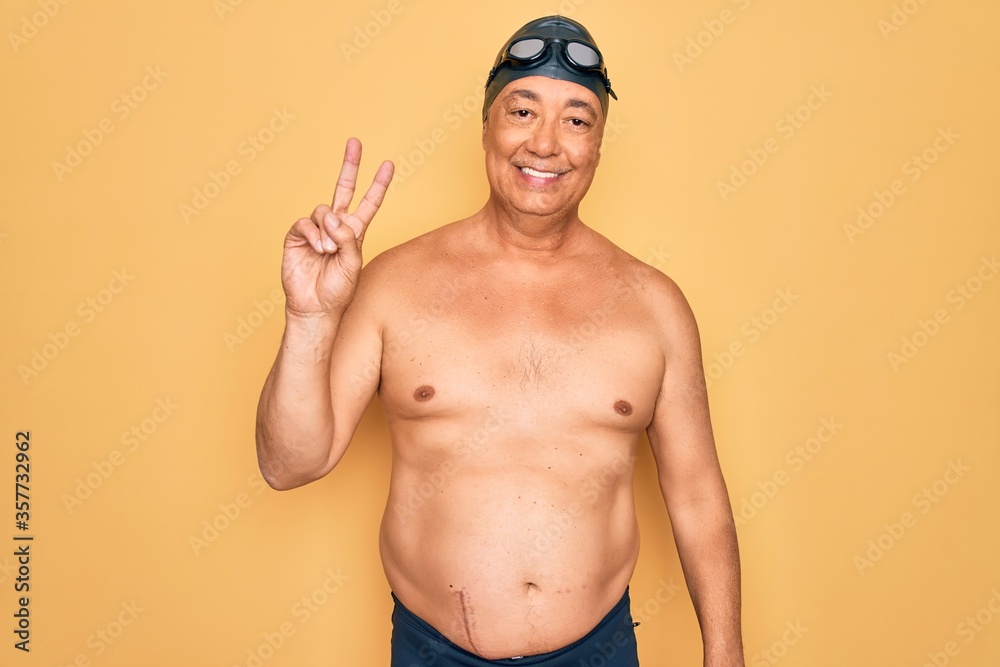Middle age senior grey-haired swimmer man wearing swimsuit, cap and goggles smiling looking to the camera showing fingers doing victory sign. Number two.