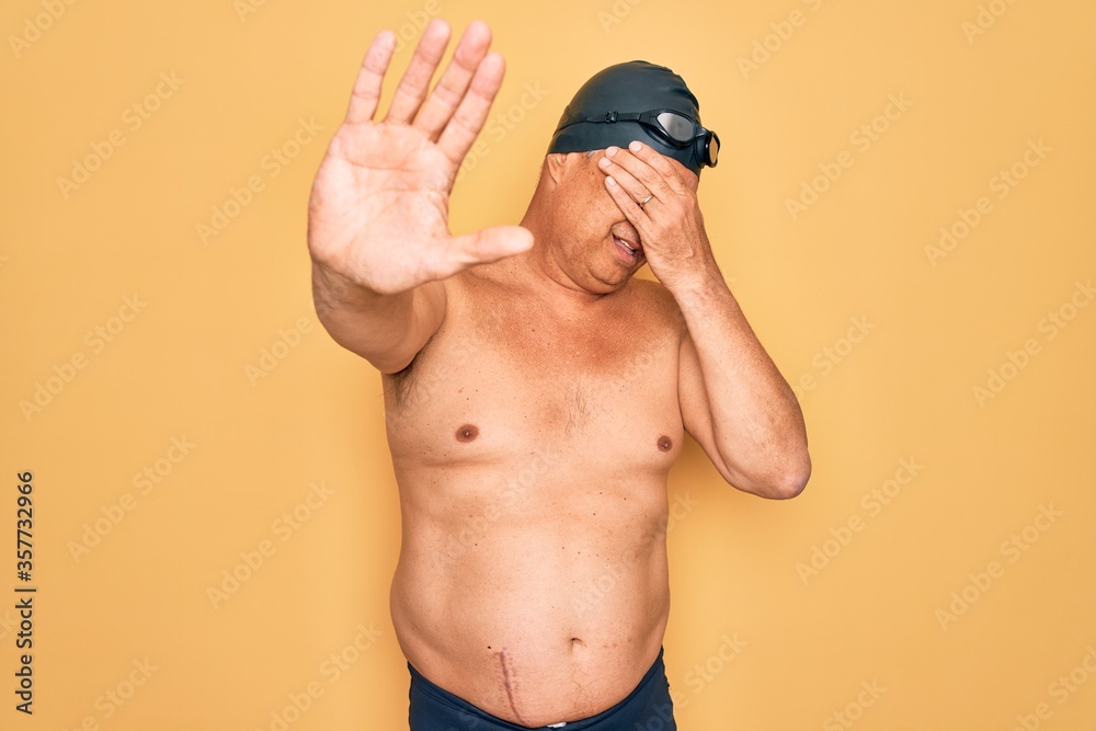 Middle age senior grey-haired swimmer man wearing swimsuit, cap and goggles covering eyes with hands and doing stop gesture with sad and fear expression. Embarrassed and negative concept.
