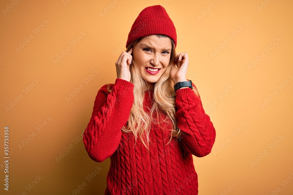 Young beautiful blonde woman wearing casual sweater and wool cap over white background covering ears with fingers with annoyed expression for the noise of loud music. Deaf concept. Young beautiful blonde woman wearing casual sweater and wool cap over white background covering ears with fingers with annoyed expression for the noise of loud music. Deaf concept.
