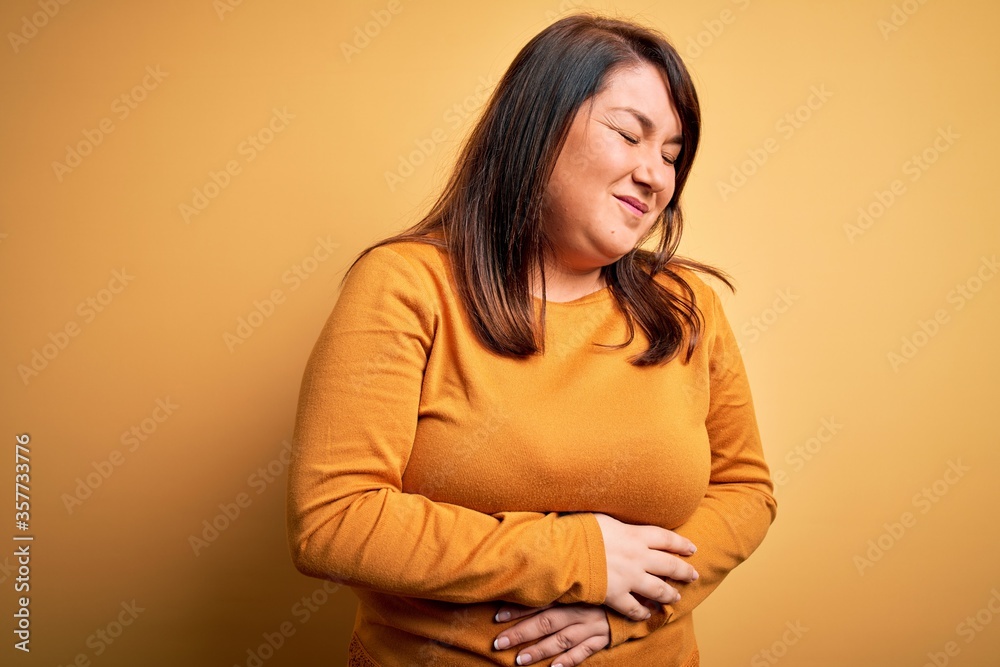 Beautiful brunette plus size woman wearing casual sweater over isolated yellow background with hand on stomach because indigestion, painful illness feeling unwell. Ache concept. Beautiful brunette plus size woman wearing casual sweater over isolated yellow background with hand on stomach because indigestion, painful illness feeling unwell. Ache concept.