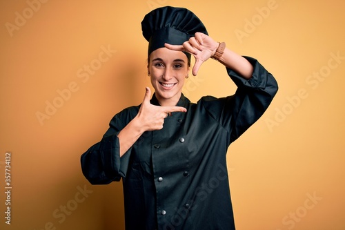 Young beautiful chef woman wearing cooker uniform and hat standing over yellow background smiling making frame with hands and fingers with happy face. Creativity and photography concept. © Krakenimages.com