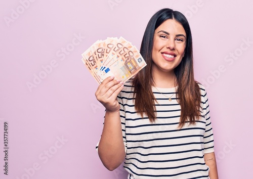 Young beautiful brunette woman holding euros banknotes over isolated pink background looking positive and happy standing and smiling with a confident smile showing teeth