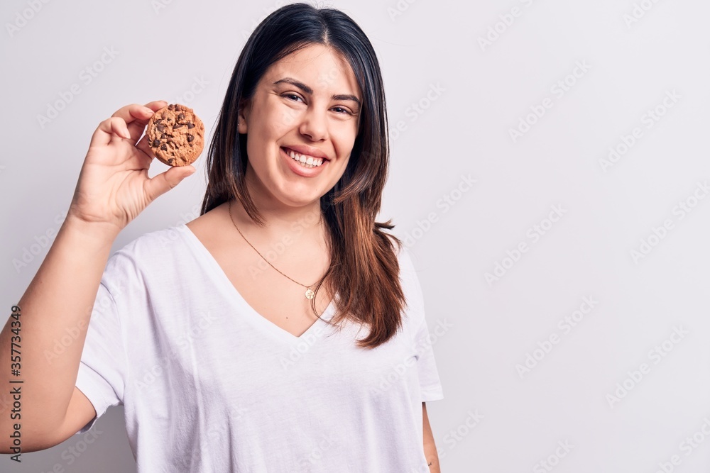Young beautiful brunette woman eating chocolate cookie over isolated white background looking positive and happy standing and smiling with a confident smile showing teeth Young beautiful brunette woman eating chocolate cookie over isolated white background looking positive and happy standing and smiling with a confident smile showing teeth