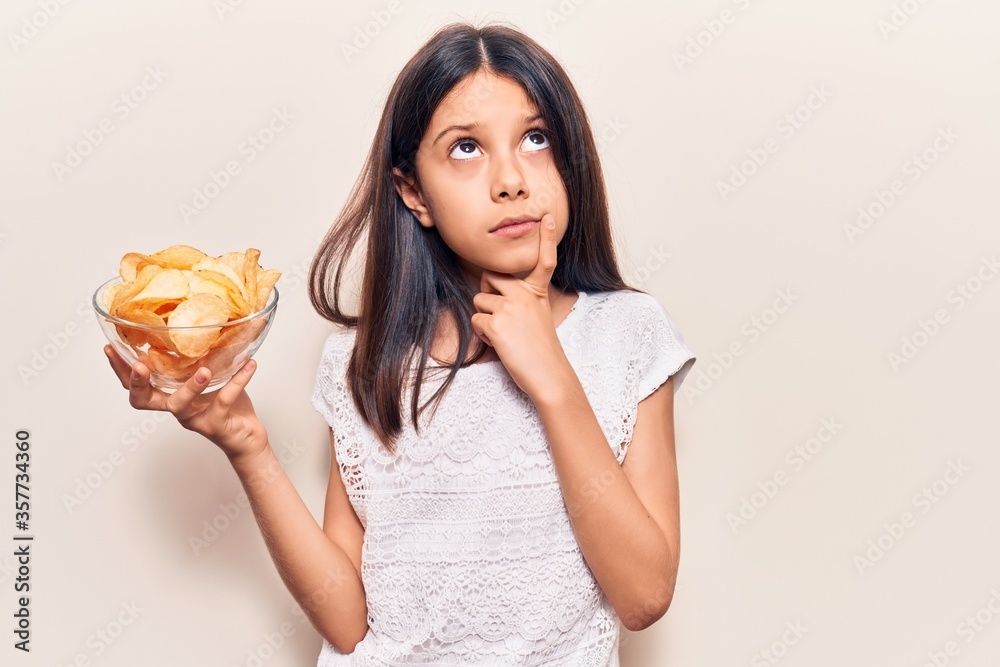 Beautiful child girl holding potato chip serious face thinking about question with hand on chin, thoughtful about confusing idea Beautiful child girl holding potato chip serious face thinking about question with hand on chin, thoughtful about confusing idea