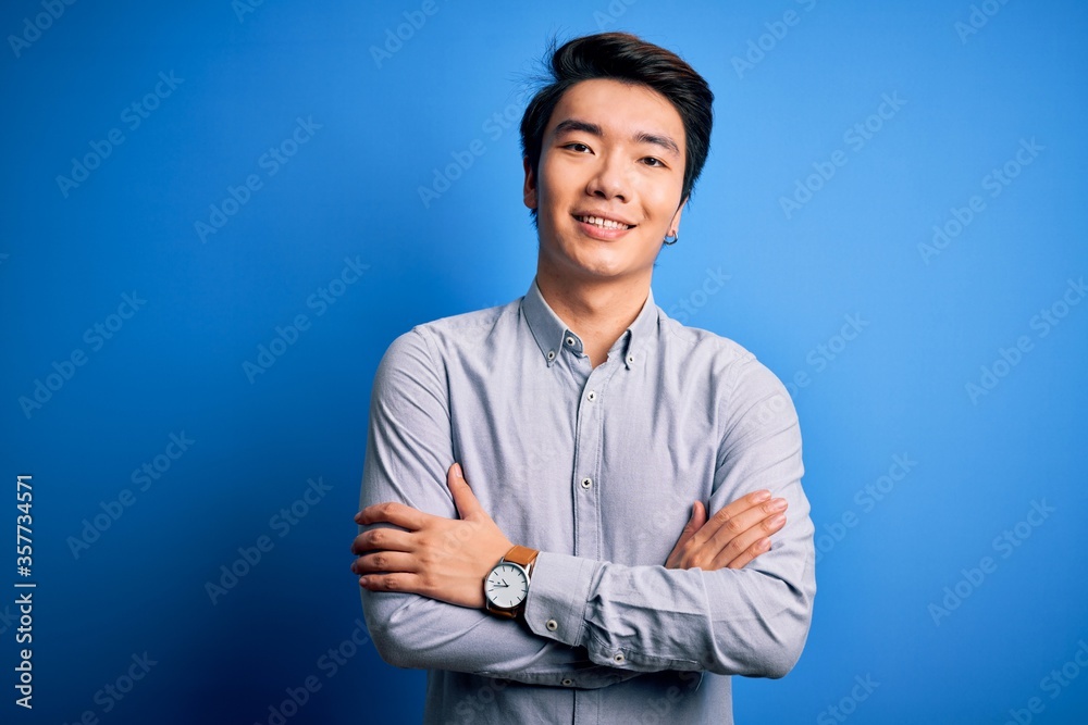 Young handsome chinese man wearing casual shirt standing over isolated blue background happy face smiling with crossed arms looking at the camera. Positive person. Young handsome chinese man wearing casual shirt standing over isolated blue background happy face smiling with crossed arms looking at the camera. Positive person.
