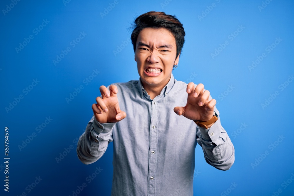 Young handsome chinese man wearing casual shirt standing over isolated blue background smiling funny doing claw gesture as cat, aggressive and sexy expression Young handsome chinese man wearing casual shirt standing over isolated blue background smiling funny doing claw gesture as cat, aggressive and sexy expression