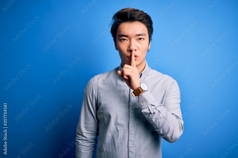 Young handsome chinese man wearing casual shirt standing over isolated blue background asking to be quiet with finger on lips. Silence and secret concept. Young handsome chinese man wearing casual shirt standing over isolated blue background asking to be quiet with finger on lips. Silence and secret concept.