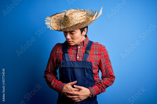 Young handsome chinese farmer man wearing apron and straw hat over blue background with hand on stomach because indigestion, painful illness feeling unwell. Ache concept.