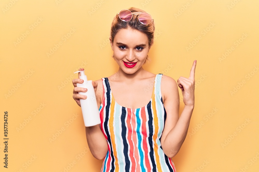 Young beautiful blonde woman wearing swimwear holding sunscreen lotion smiling happy pointing with hand and finger to the side Young beautiful blonde woman wearing swimwear holding sunscreen lotion smiling happy pointing with hand and finger to the side