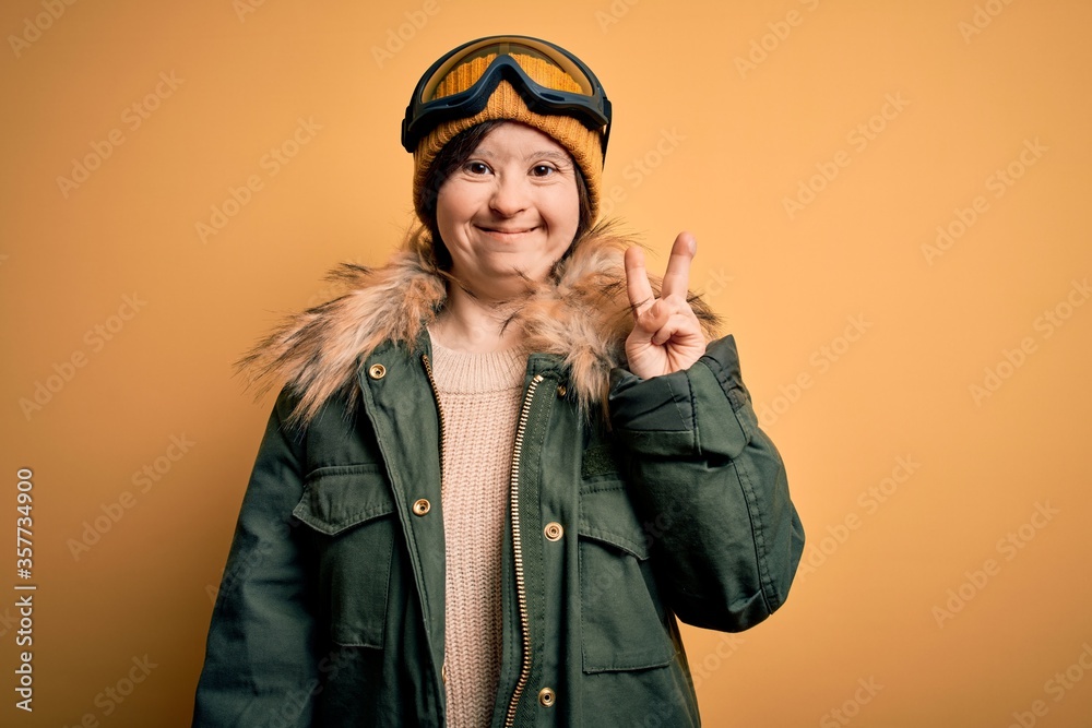 Young down syndrome woman wearing ski coat and glasses for winter weather smiling with happy face winking at the camera doing victory sign. Number two.