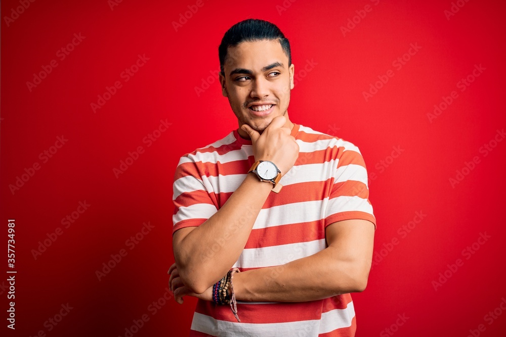 Young brazilian man wearing casual striped t-shirt standing over isolated red background with hand on chin thinking about question, pensive expression. Smiling with thoughtful face. Doubt concept. Young brazilian man wearing casual striped t-shirt standing over isolated red background with hand on chin thinking about question, pensive expression. Smiling with thoughtful face. Doubt concept.