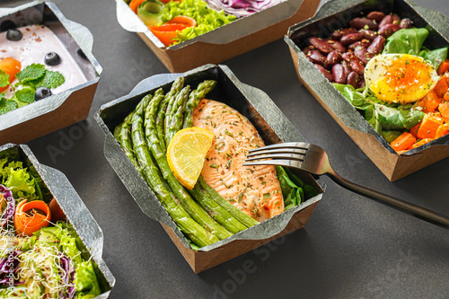 Ready healthy food catering menu in lunch boxes fish and vegetable packages as daily meal diet plan courier delivery with fork isolated on black table background. Take away containers order concept. photo