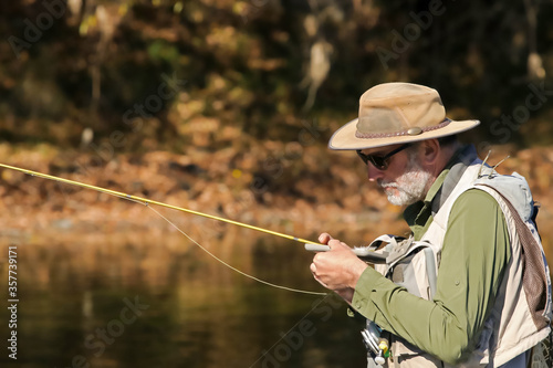 Mature bearded man choosing a fly from his fly box and holding his fishing rod amidst autumn colors © Susan