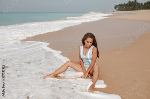 Sensual young brunette beauty wearing colorful stripped swimsuit  sitting on the beach over beautiful sea, sky and tropical island background, waves touch her feet © Yevhenii