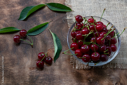 red cherries in a transparent glass bowl on a wooden background, with green leaves of a cherry tree. Summer berries. Yummy.