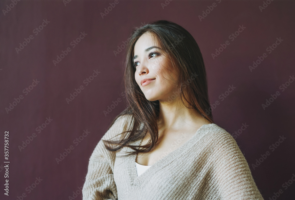 Charming smiling asian girl with long brown hair in beige sweater looking at camera isolated on dark bordo background Charming smiling asian girl with long brown hair in beige sweater looking at camera isolated on dark bordo background