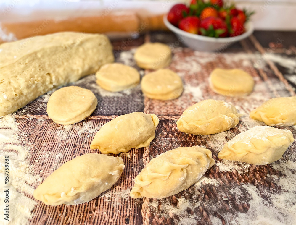 Uncooked dumplings with strawberries and dough on an abstract wooden surface. The process of preparing a traditional meal of flour dough.