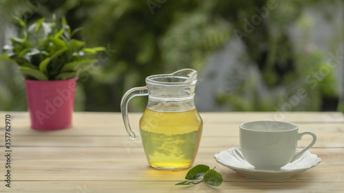 Green tea jar with tea leaves and cup of coffee on wood board and blurred green leaves in pink tin background