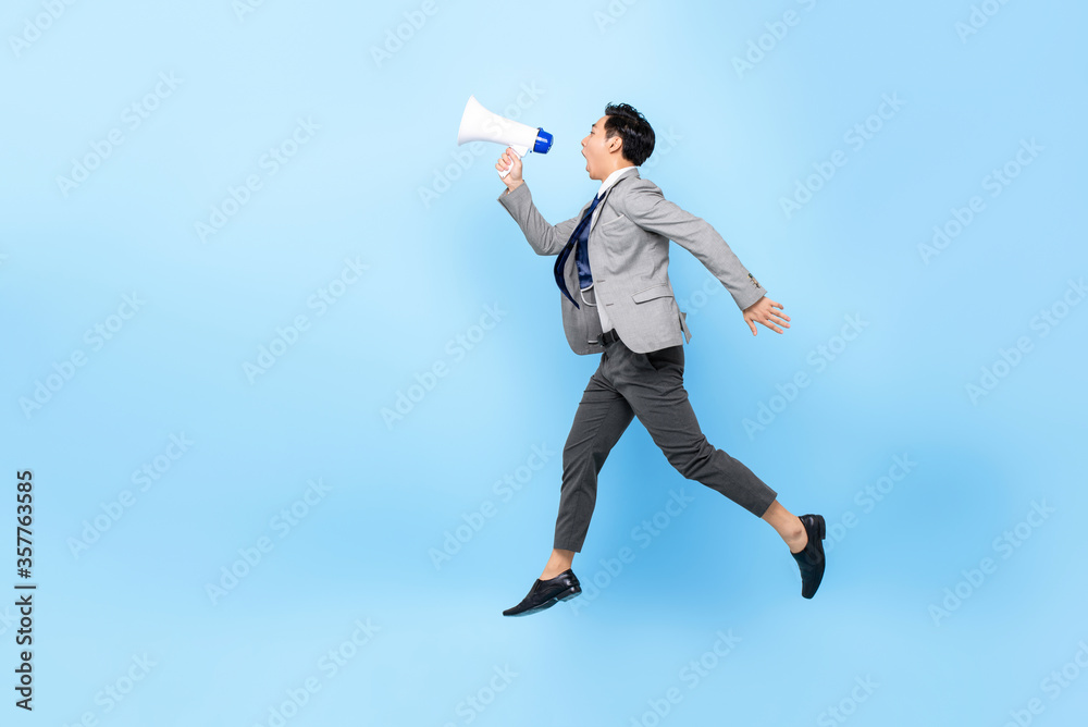 Young Asian businessman jumping and shouting on megaphone isolated on light blue background