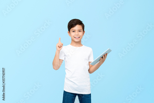 Cute 10 year-old mixed race boy holding tablet computer with one hand pointing to empty space upward isolated on light blue background
