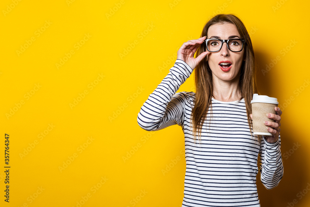 Young woman shocked in glasses with a paper cup of coffee on a yellow background Young woman shocked in glasses with a paper cup of coffee on a yellow background