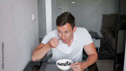 sporty muscular guy in white t-shirt eats delicious porridge cereals with blueberries and milk for breakfast in hotel restaurant