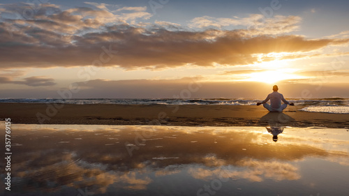 yoga meditation silhouette lotus sunrise beach, mindfulness, wellness and wellbeing concept, water reflection of man in yoga lotus pose sitting alone on sand with ocean cloud background, copy space