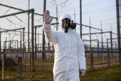 Researcher wearing white protective uniform, gas mask and gloves on the territory of thermal power station, shows stop sign, power lines on background