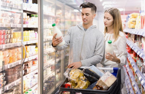 Young smiling female and male customers choosing milk and dairy products in grocery, family shopping