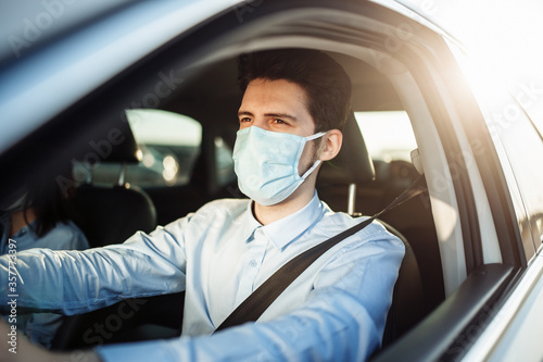 Young boy taxi driver gives passenger a ride wearing sterile medical mask. A man in the car behind the steering wheel works during coronavirus pandemic. Social distance and health safety concept. © Konstantin Zibert