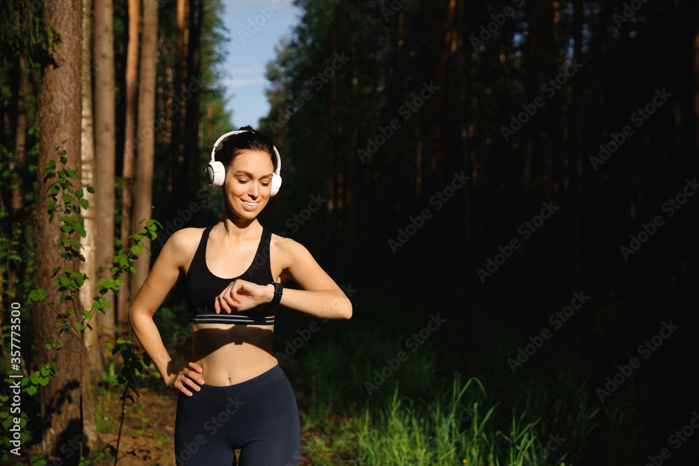 A beautiful Girl in leggings, t-shirt and white Bluetooth headphones stands in the woods after a run and looks at her smart watch A beautiful Girl in leggings, t-shirt and white Bluetooth headphones stands in the woods after a run and looks at her smart watch