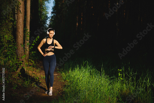 A beautiful Girl in leggings, t-shirt and white Bluetooth headphones stands in the woods after a run and looks at her smart watch