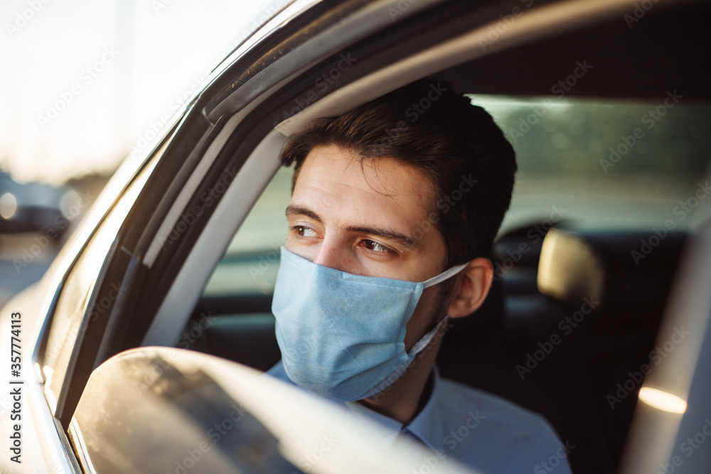 Young businessman takes a taxi and looks out of the car window wearing sterile medical mask. A man sits on the back seat of taxi and takes a ride during coronavirus pandemic. Social distance concept. Young businessman takes a taxi and looks out of the car window wearing sterile medical mask. A man sits on the back seat of taxi and takes a ride during coronavirus pandemic. Social distance concept.