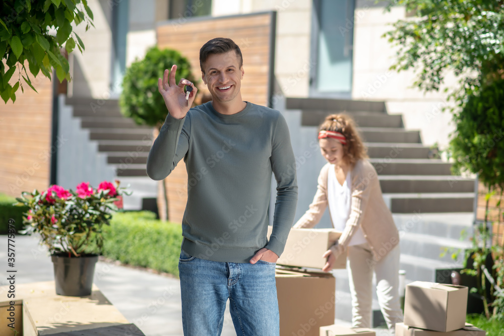 Naklejka premium Smiling man holding a key from the new house and feeling great, his wife standing near the cardboards