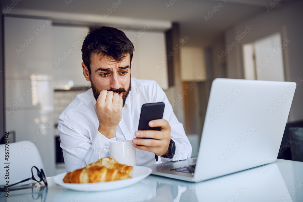 Young attractive bearded man getting nervous about message from his boss. he is biting his nails. Morning time, dinning room interior. Young attractive bearded man getting nervous about message from his boss. he is biting his nails. Morning time, dinning room interior.