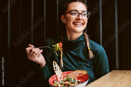 Canvas Print Young attractive teenage girl with healthy habits sitting in restaurant, holding bowl with salad and having lunch